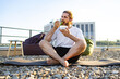 © sofiko14 - A man with a beard and tied-back hair sits cross-legged on a yoga mat on a gravel rooftop, eating a salad from a plastic container