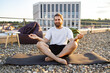 © sofiko14 - A bearded man in casual attire practices yoga and meditation on a gravel rooftop with a city skyline in the background during golden hour