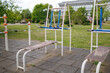 © WoodHunt - Outdoor workout area with blue and white metal exercise machines on a paved rubber tile ground. Trees and a modern building are in the background under a cloudy sky