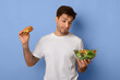© Prostock-studio - A person stands with a burger in one hand and a salad bowl in the other. The background is blue, and the person shows uncertainty about their food choice.