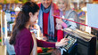 © Prostock-studio - A worker at a grocery store scans items for customers at the register. Two shoppers stand nearby, waiting for their turn. The setting shows a busy store with various products around.