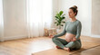 © Irina - Pregnant woman practicing yoga meditation on mat in bright room. Pregnant woman sits cross-legged in serene pose, surrounded by natural light and indoor plant.