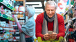 © Prostock-studio - A man stands in a grocery store looking at his smartphone while pushing a shopping cart. A woman browses shelves in the background. The store is well-lit and busy with products.