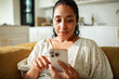 © Marko Geber - Smiling woman using smartphone on sofa at home