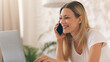 © Prostock-studio - A woman sits at a table with a laptop in front of her. She is smiling while talking on the phone. The room is bright and has a plant in the background.