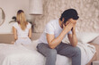 © Prostock-studio - A man sits on the edge of a bed with his hands covering his face. A woman sits in the background, facing away. They appear to be experiencing a difficult moment in their relationship.