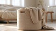 © CYBERPINK - Beige-colored storage basket with a handle on top. the basket is placed on a wooden floor in a bedroom with a bed in the background.