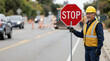 © OlgaNeuroArt - Male traffic control worker holding a stop sign on a road. Senior man in safety vest and hard hat at construction site. Professional flagger directing traffic
