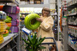 © Ivan Traimak - Woman shopping for garden supplies, selecting a green hose while pushing a trolley