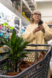 © Ivan Traimak - Woman standing with a shopping cart in an aisle, looking up while holding a smartphone