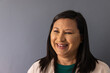 © wavebreak3 - Mature woman smiling and posing against gray wall wearing green top beige jacket and stud earrings