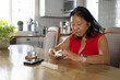 © Wavebreak Media - Senior Asian woman in red blouse examining device at kitchen table with pill tray, copy space