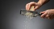 © Lee - Person's hand grating a block of hard cheese with a metal grater, creating shredded cheese for cooking.