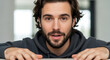 © Natalia - Close up portrait of young bearded man leaning forward looking at camera with surprised expression in gray hoodie modern indoor setting natural lighting authentic human emotion realistic photography.