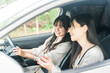 © buritora - A woman driving a car and another woman in the passenger seat searching on her smartphone.