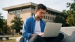 © Aqeela - Young man using a modern laptop while sitting outside on a campus university stone bench