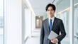 © YT - 明るく清潔感のあるオフィス廊下でノートパソコンを抱えて立つ若いビジネスマンのポートレート。スーツ姿と落ち着いた表情が信頼感と知性を印象づける、採用、企業案内、営業、キャリア、ビジネスコミュニケーションに適した現代的な人物イメージ
