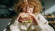 © liliyabatyrova - Bright sunlight bathes a young individual with curly hair as they prepare to take a big bite of a delicious sandwich. The cozy indoor atmosphere invites joy and comfort