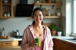 © miss irine - Woman smiles holding green smoothie in kitchen. Lady drinks healthy beverage after workout, feeling energised. She wears sporty clothes at home.