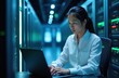 © miss irine - Young asian woman works on laptop in data center server room. Rows of glowing racks with computer equipment are visible. She focuses on screen, typing. Tech career woman studies server diagnostics.