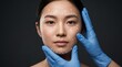 © Olga Ко - Plastic surgery and facial contouring concept. Portrait of a beautiful Asian woman with white surgical guidelines on her face, touched by doctor hands in blue gloves against a dark background