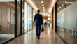 © Vhagar - A businessman in a suit walks through a modern office hallway, carrying a briefcase. The setting features glass partitions and contemporary lighting.