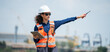 © winnievinzence - Female site engineer in a hard hat and safety vest uses a tablet and radio to direct logistics operations at a busy industrial shipping port