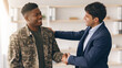 © Prostock-studio - Two men are standing in a modern office space. One is wearing a military jacket. They are smiling and shaking hands, indicating a positive interaction.