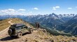 © Mey - Two hikers stand on a rocky mountain peak, admiring a panoramic view of a vast mountain range. A green Jeep Wrangler sits atop the peak, equipped with a rooftop cargo rack.