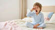 © OlgaNeuroArt - Young boy in striped pajamas yawning and rubbing his eyes on a bed. Sleepy child waking up in the morning. Bedtime and morning routine concept