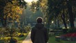 © pisan thailand - A person in a dark jacket walks alone down a sunlit park path surrounded by trees and greenery in the late afternoon.