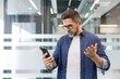 © Liubomir - Mature professional holding smartphone, looking frustrated and confused after reading bad news or an upsetting message in a modern office environment, stressed and pensive