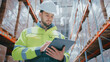 © ihorvsn - Bearded Caucasian man in high-visibility jacket and helmet using digital tablet while standing near tall racks in warehouse. Reviewing stock data or updating inventory information during inspection.