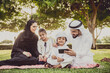 © oneinchpunch - Arabian family in traditional dress smiling together on a picnic blanket in a sunny green park, parents and children enjoying leisure, bonding and relaxation outdoors