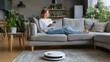 © standret - Young woman using smartphone while relaxing on gray sofa in modern living room with indoor plants and robotic vacuum cleaner on wooden floor