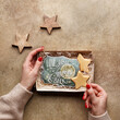 © Zygonema - Female hands in sweater holding cardboard gift box with decorated gingerbread cookies in shape of stars and with 'Love you to the moon and back' text over beige concrete background. Overhead view