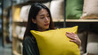 © A2Z AI  - Young woman thoughtfully selects soft yellow cushion, eyes closed, enjoying the texture in a home decor store aisle with blurred merchandise racks