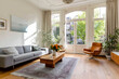 © KAHAR - A sunlit, airy living room with large windows, neutral tones, wood floors, and touches of color in furniture and art