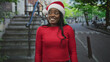 © Krakenimages.com - Woman wearing santa hat smiling with a visible grin on a street in front of building stairs, red sweater visible; holiday joy.