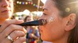 © lynkid1966 - A woman's face is being painted with glitter by another person during a lively summer festival event outdoors.