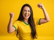 © Iwan - Excited young Brazilian woman fan with face paint cheering and celebrating a victory with fists raised on a yellow background.