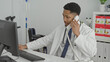 © Krakenimages.com - Man holding phone to ear while typing on computer at a clinic desk, stethoscope visible; focused care.