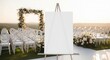 © Nataly - Elegant wedding setup with blank canvas on easel in outdoor venue. Wedding ceremony features white chairs and floral decorations creating romantic ambiance.