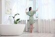 © New Africa - Woman in bathrobe opening beautiful window curtains in bathroom