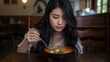 © VolumeThings - Young Asian woman with long dark hair in casual attire blowing on a bowl of traditional soup at a local eatery during lunch.