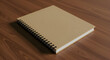 © joe - A neutral-toned spiral-bound notebook rests on a wooden surface. A blank, kraft-colored notebook sits atop a smooth, light brown wooden surface.
