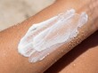 © Marian - Primer plano de crema protectora solar mineral de zinc aplicándose en un brazo bronceado con arena y cristales de sal marina.