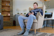© Marina Demidiuk - Handsome young man using social media over smart phone against living room background. Bearded guy looking at phone while sitting on chair at home. Full length photo. Young adult gen y use hold mobile
