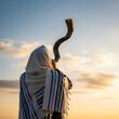 © we.bond.png - A religious man wearing a tallit shawl blows a traditional shofar horn during a peaceful and golden sunset outdoor session