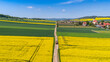 © Adam - Wide aerial drone view of a farm tractor driving on a dirt road that divides vibrant yellow blooming rapeseed fields and green crops, with a village, church, and hills in the distance.